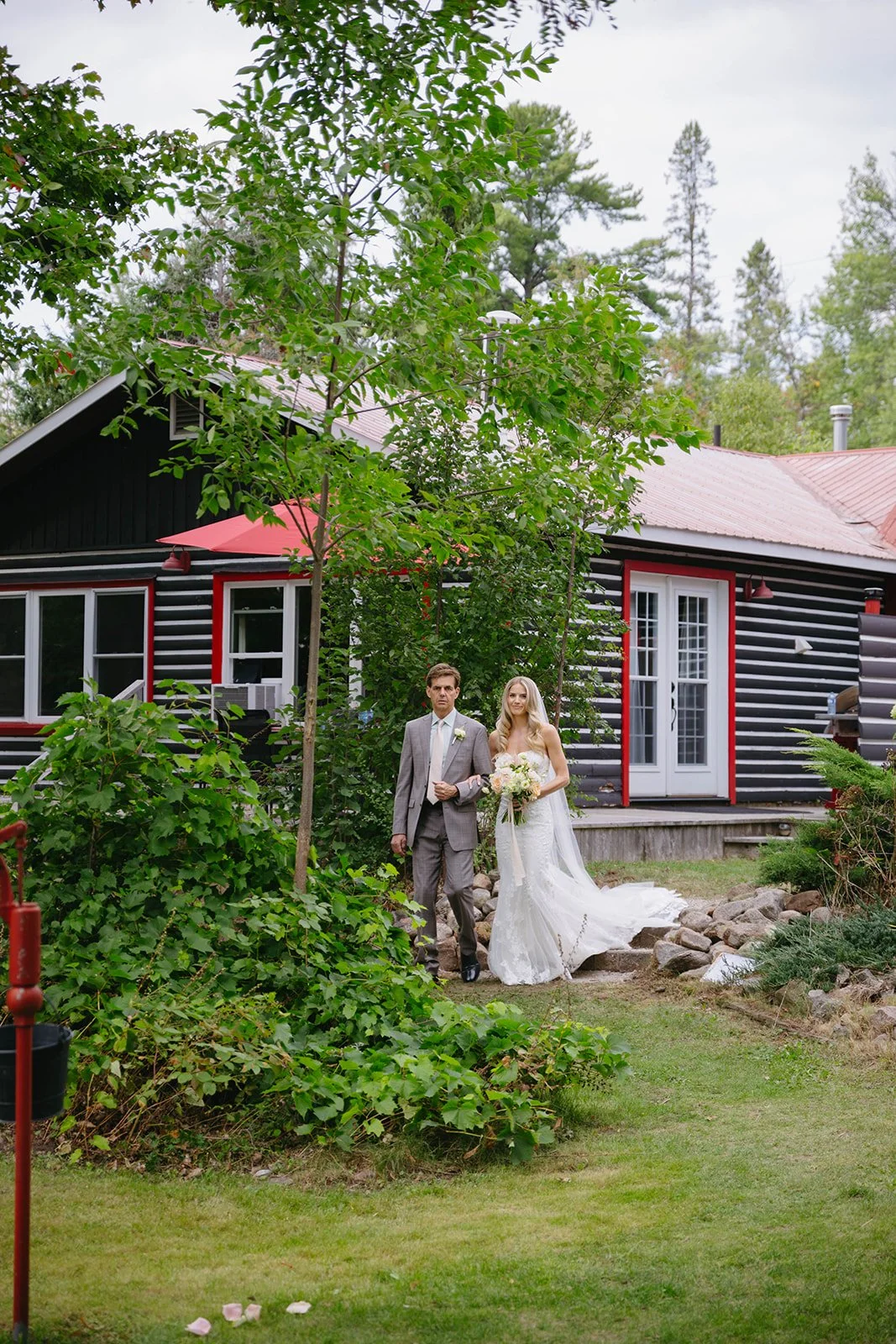 A bride in a white wedding dress holding a bouquet of flowers walks with a groom in a gray suit outdoors in front of a black house with red and white trim. Trees and greenery surround the scene.