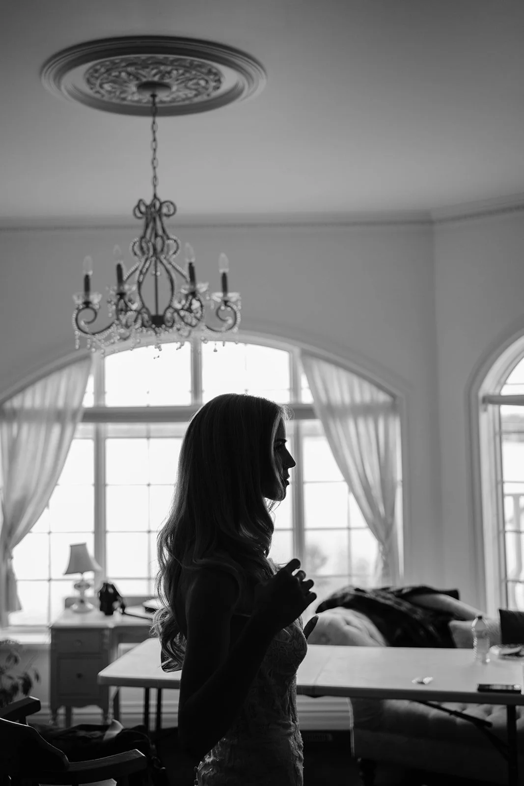 Silhouette of a woman with long hair in a room with large windows, curtains, and a chandelier.
