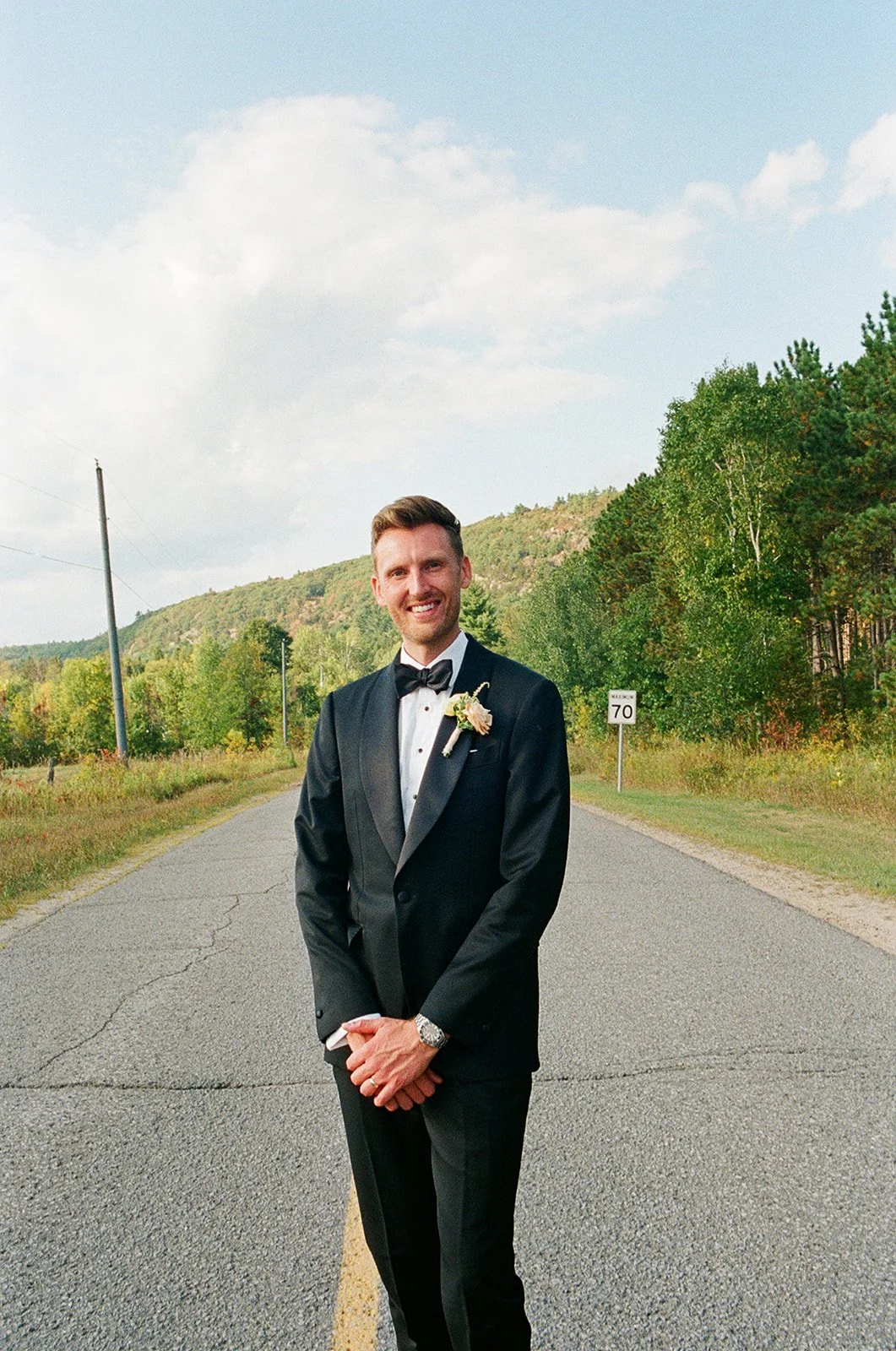 A man in a tuxedo standing on an empty rural road, smiling, with green trees and hills in the background.