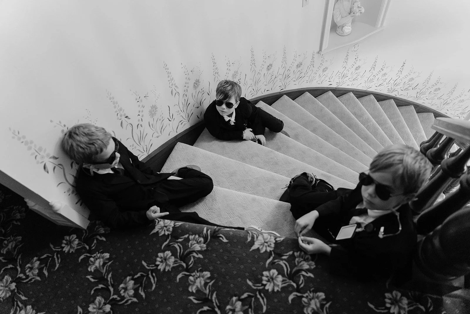 Three boys wearing sunglasses and black suits sitting on a staircase, two leaning against the wall and one sitting on the stairs, with a woman standing in the background.