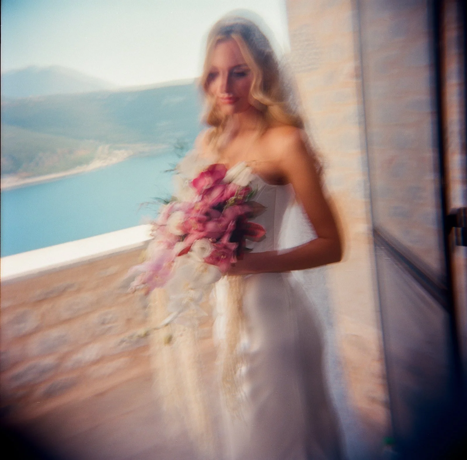 Blurry photo of a woman holding a bouquet of pink and white flowers, standing near a window with a view of a body of water and mountains in the background.