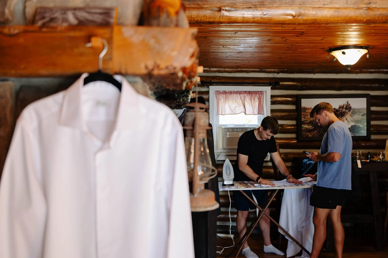A white dress shirt hanging on a hook in the foreground, with two young men in casual clothing preparing in a rustic log cabin interior in the background, with a window, framed picture, and ceiling light.