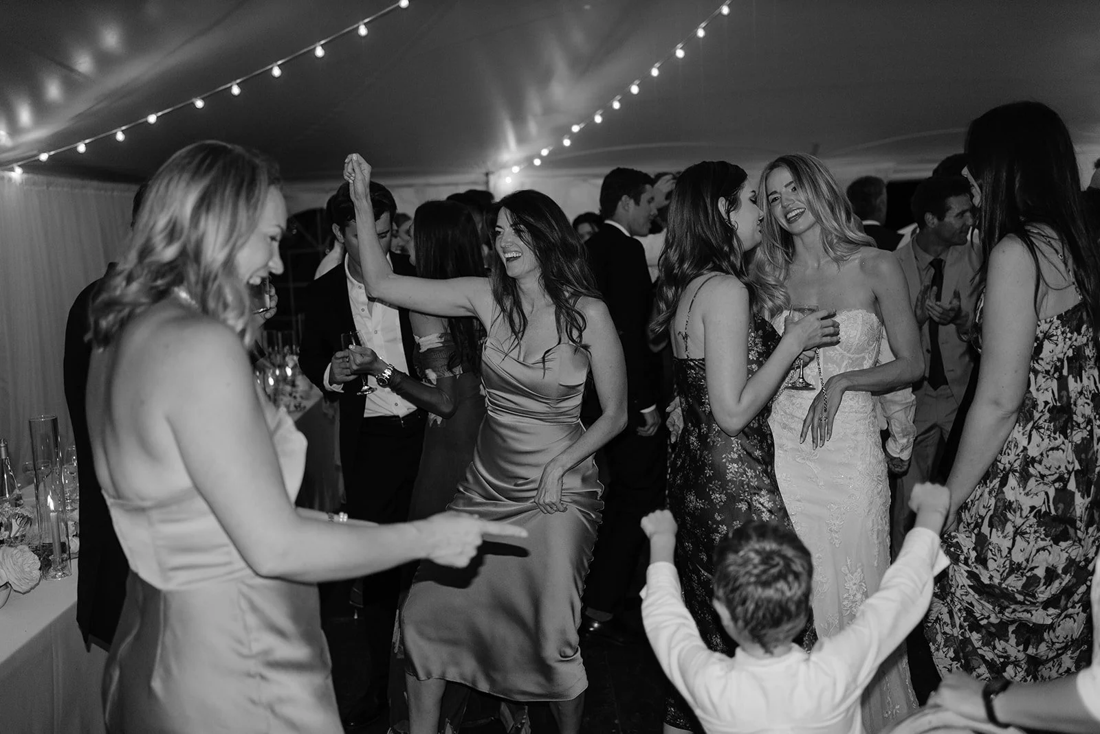 People dancing and socializing at a party or celebration in a tent with string lights