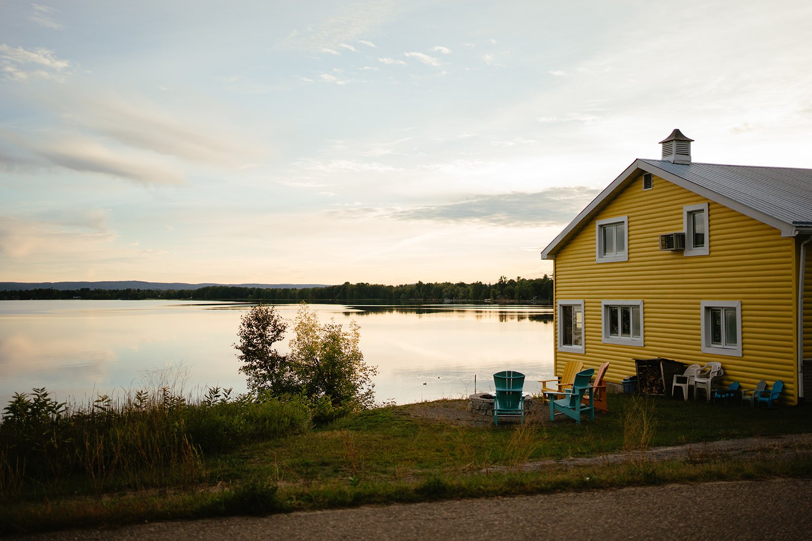 Yellow house with white-trimmed windows beside a calm lake, with colorful outdoors chairs on the lawn and trees in the background.