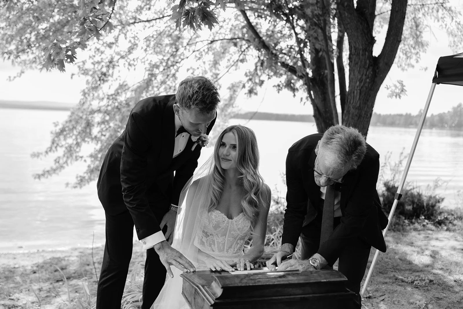 A black and white photo of a wedding ceremony outdoors by a lake, with a bride in a white dress and two men in black tuxedos signing a document under a tree.