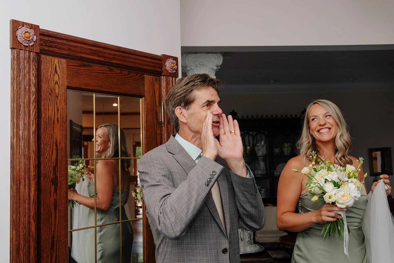 A man in a gray suit is whispering or shouting into a woman's ear as she smiles, holding a bouquet of white flowers. A mirror behind them reflects the woman's back and the man’s face.