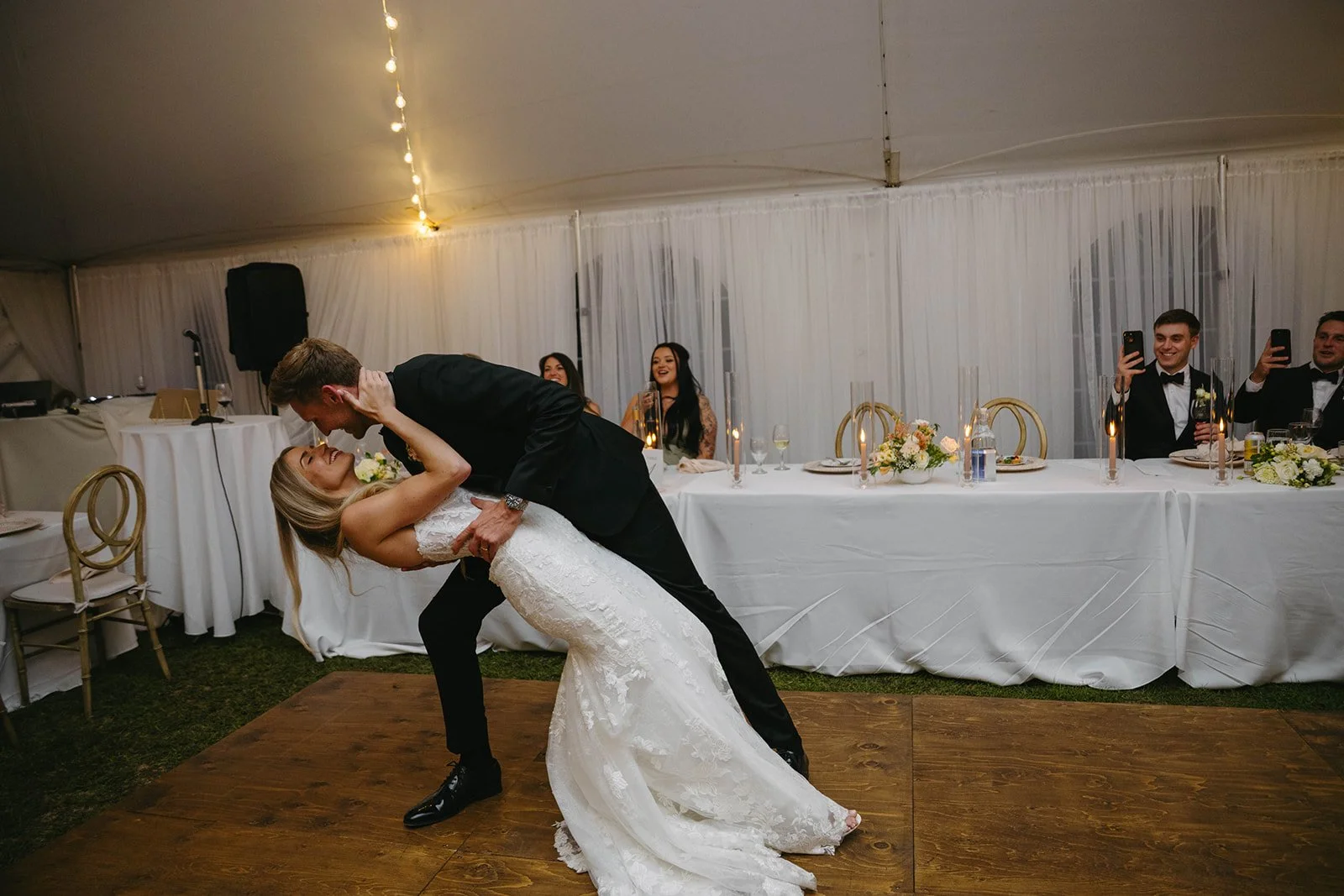 A bride and groom dancing closely at their wedding reception, with guests watching and taking photos in the background under a decorated tent.