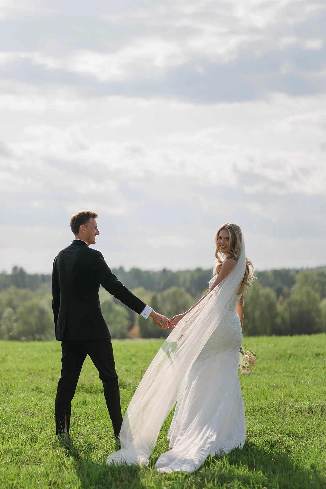 A bride and groom holding hands and smiling outdoors on a grassy field with cloudy sky and trees in the background.