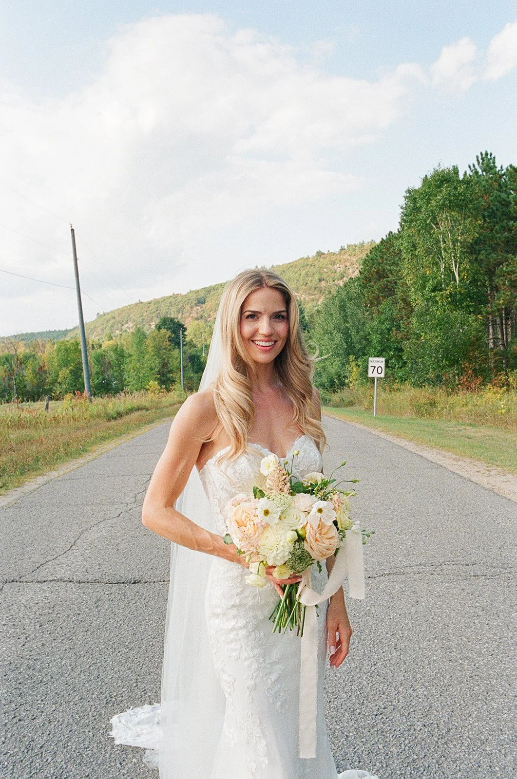 A smiling bride in a white wedding dress holding a bouquet of flowers on a rural road with trees and hills in the background.