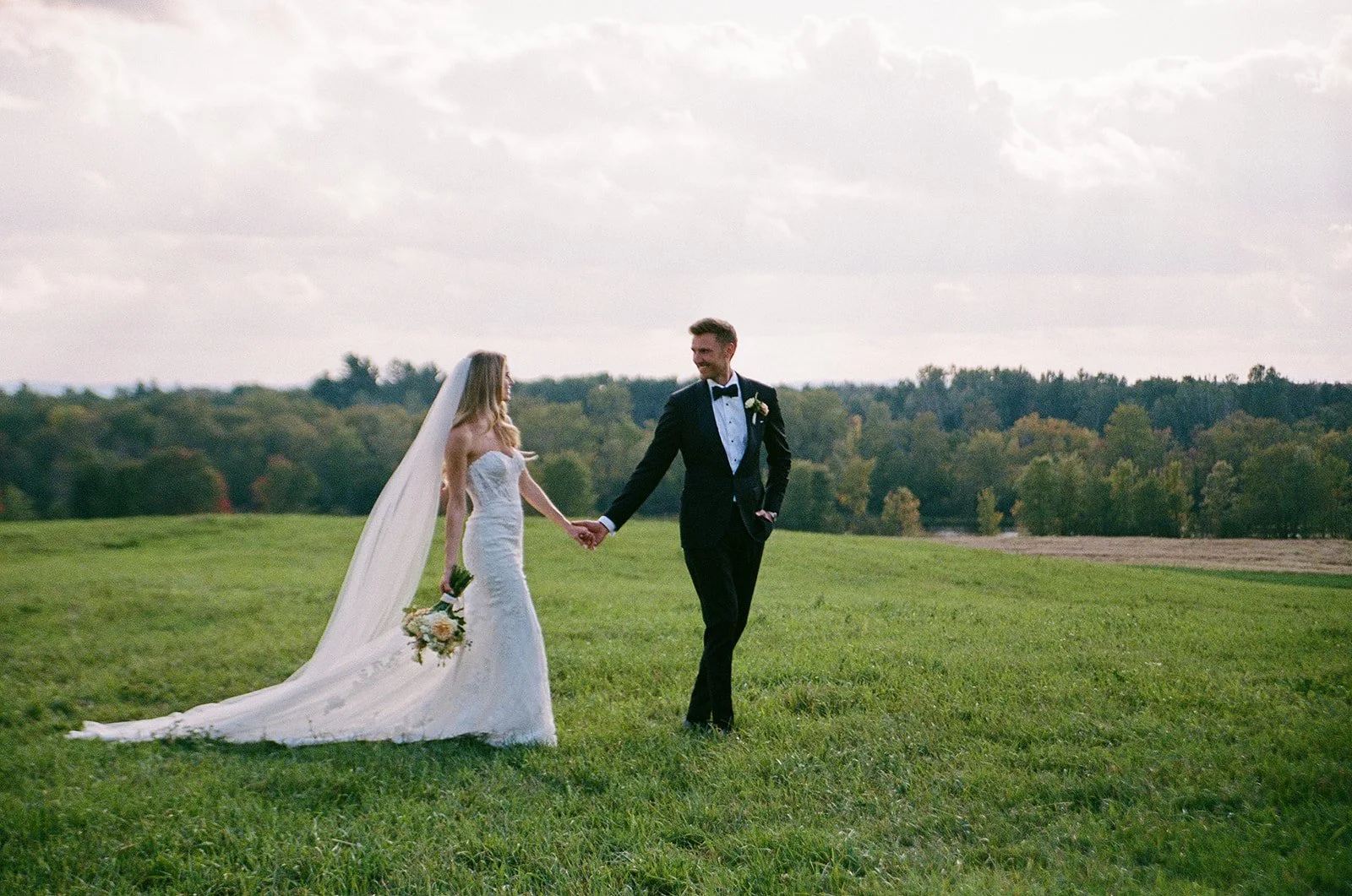 A bride and groom holding hands in a grassy field with trees in the background, dressed in wedding attire.