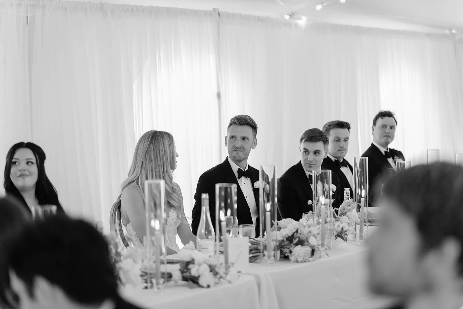 Black and white photo of a wedding reception with five people seated at a table, dressed in formal attire. The men are wearing tuxedos with bow ties, and the woman is wearing a gown. The table is decorated with floral arrangements and tall candle hol