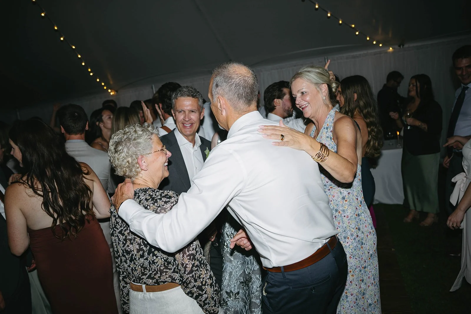 People dancing and celebrating at a wedding reception under string lights, smiling and enjoying each other's company.