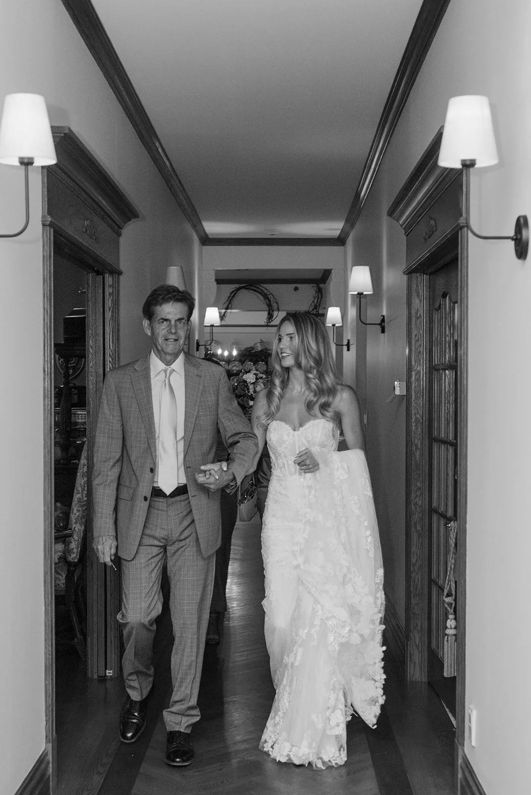 A bride walking down a hallway with her arm linked to a man, possibly her father, at her wedding, smiling and looking at him.