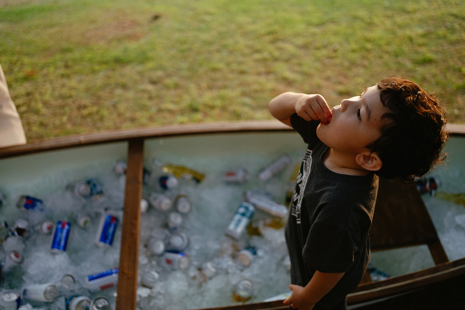 Young boy standing near a wooden boat filled with crushed cans and trash, outdoors on a grassy field.