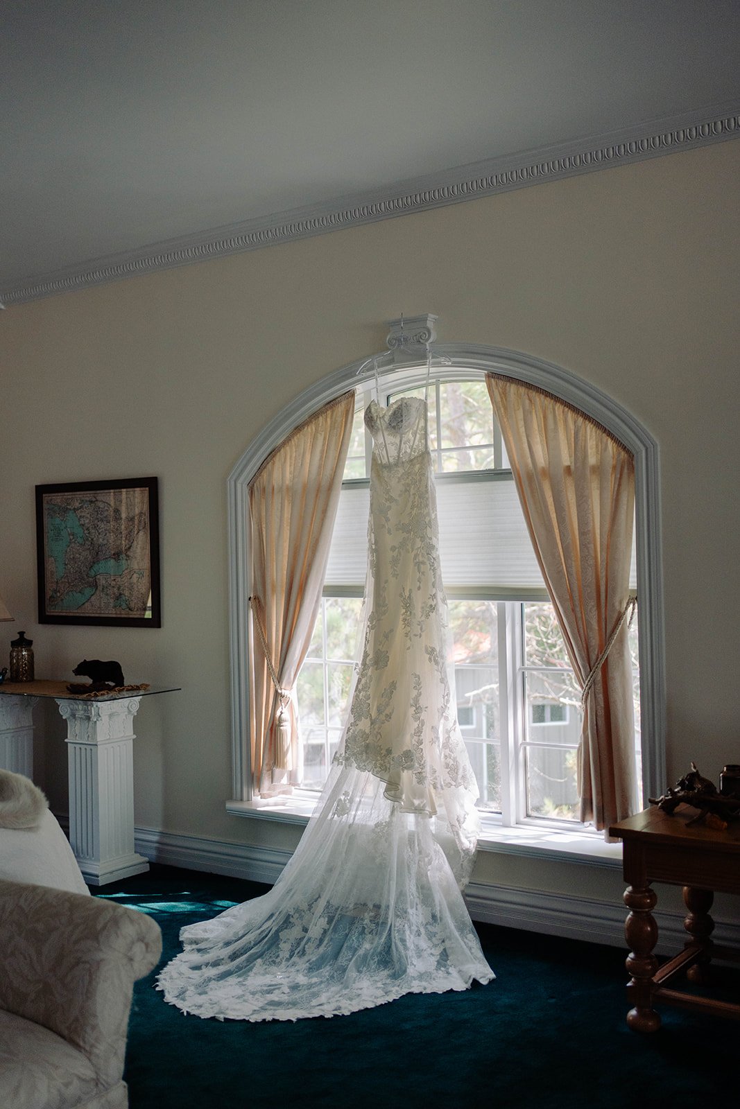 A wedding dress hanging in front of a window with curtain panels, in a well-lit room.
