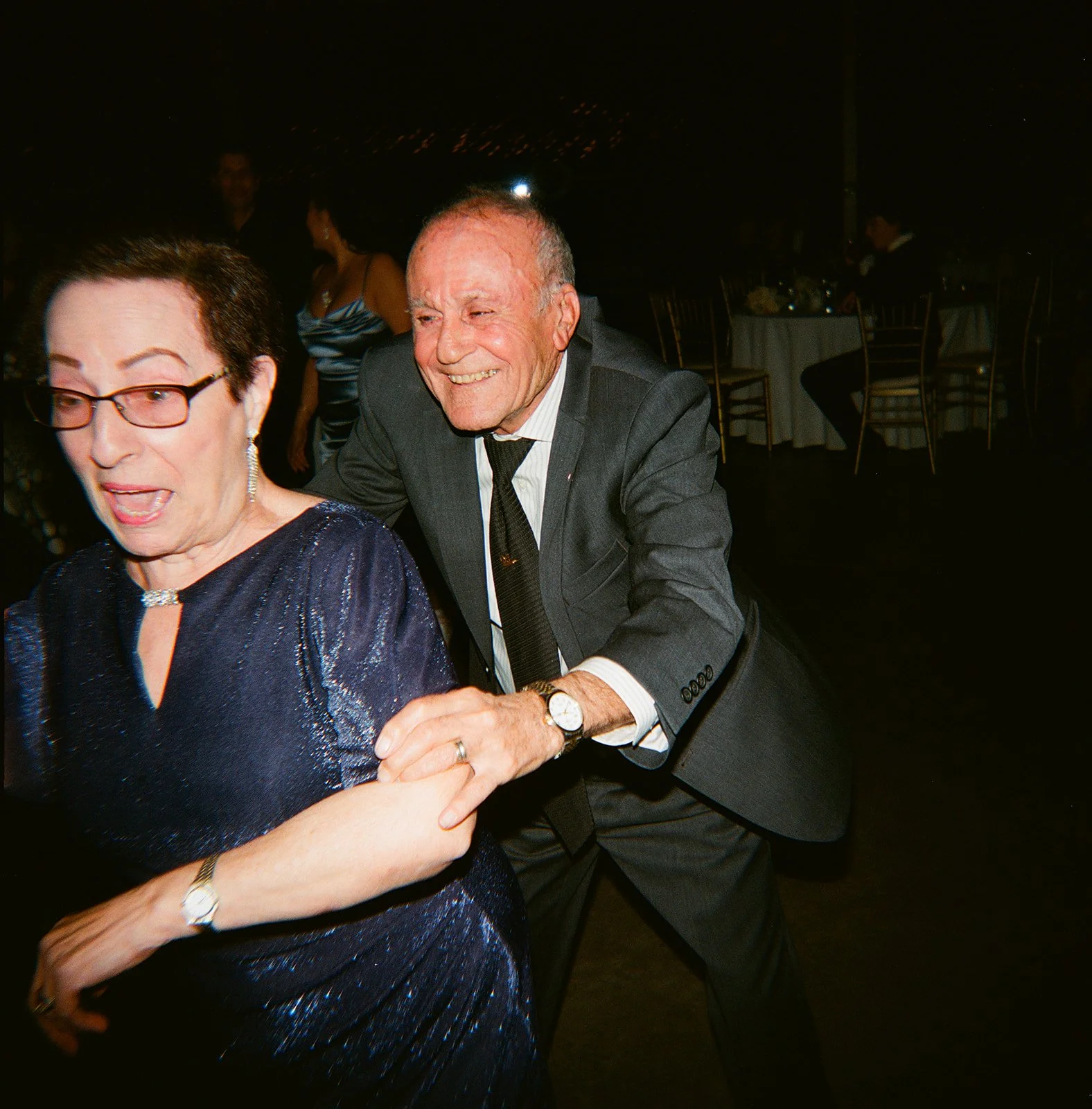 An elderly man in a gray suit and black tie playfully pulls on a woman in a dark blue, shiny dress with glasses; they are at a formal event, with round tables and chairs in the background.