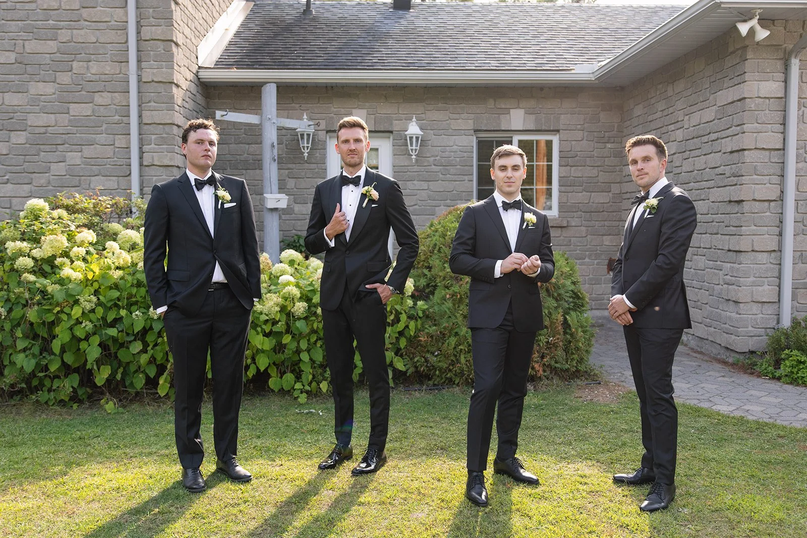 Four men in black tuxedos with white shirts and bow ties standing outdoors on a lawn in front of a brick house with lush greenery and hydrangea bushes.
