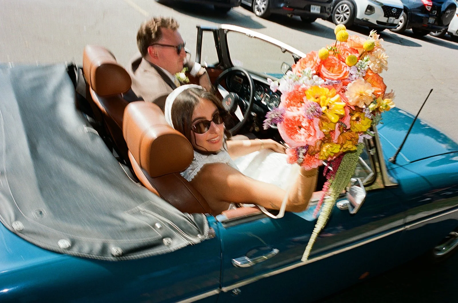 A couple dressed in wedding attire riding in a vintage blue convertible car decorated with a large colorful bouquet of flowers on the hood, surrounded by parked cars.