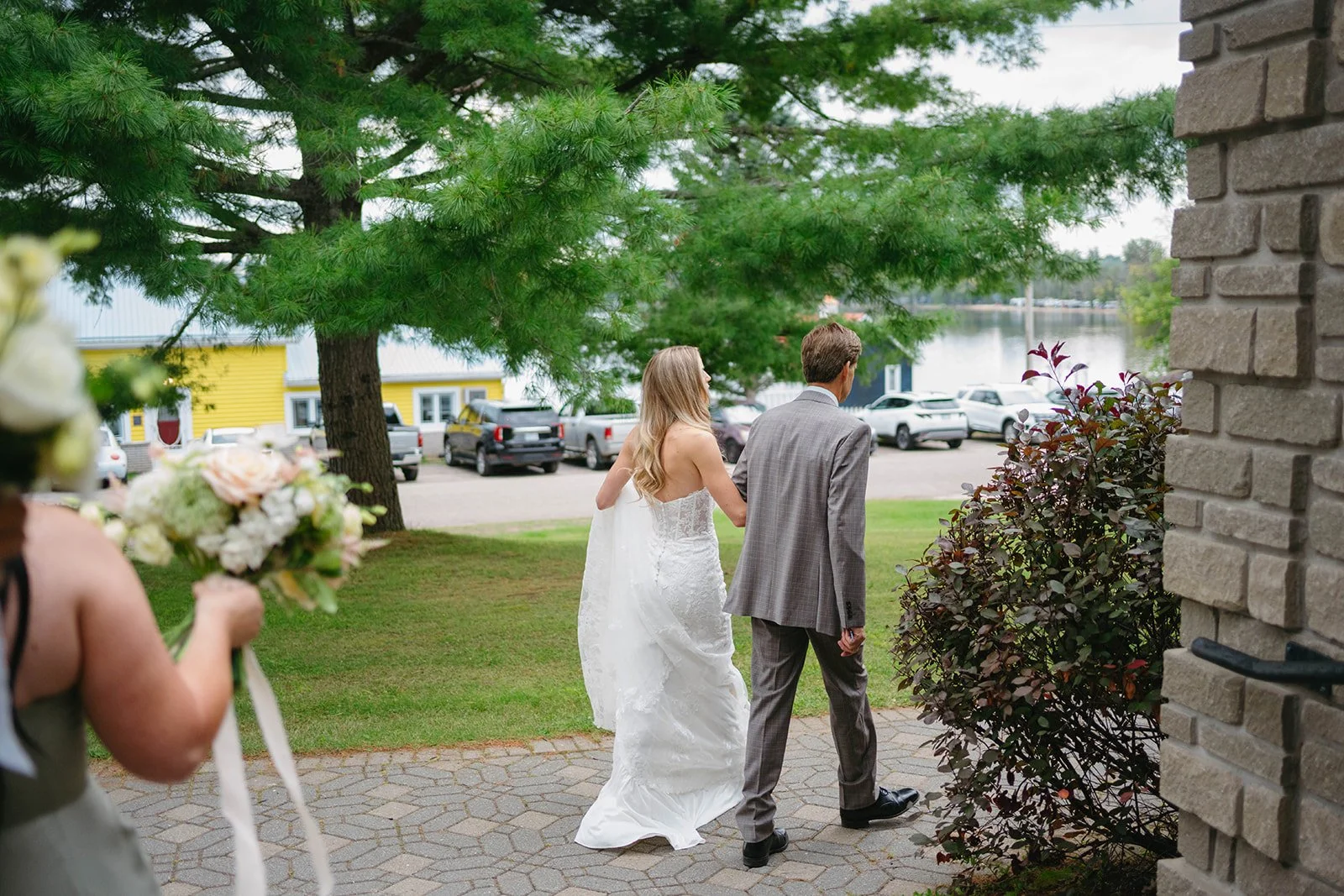 A bride and groom walking together outdoors near a lake, with a woman holding a bouquet visible in the foreground.