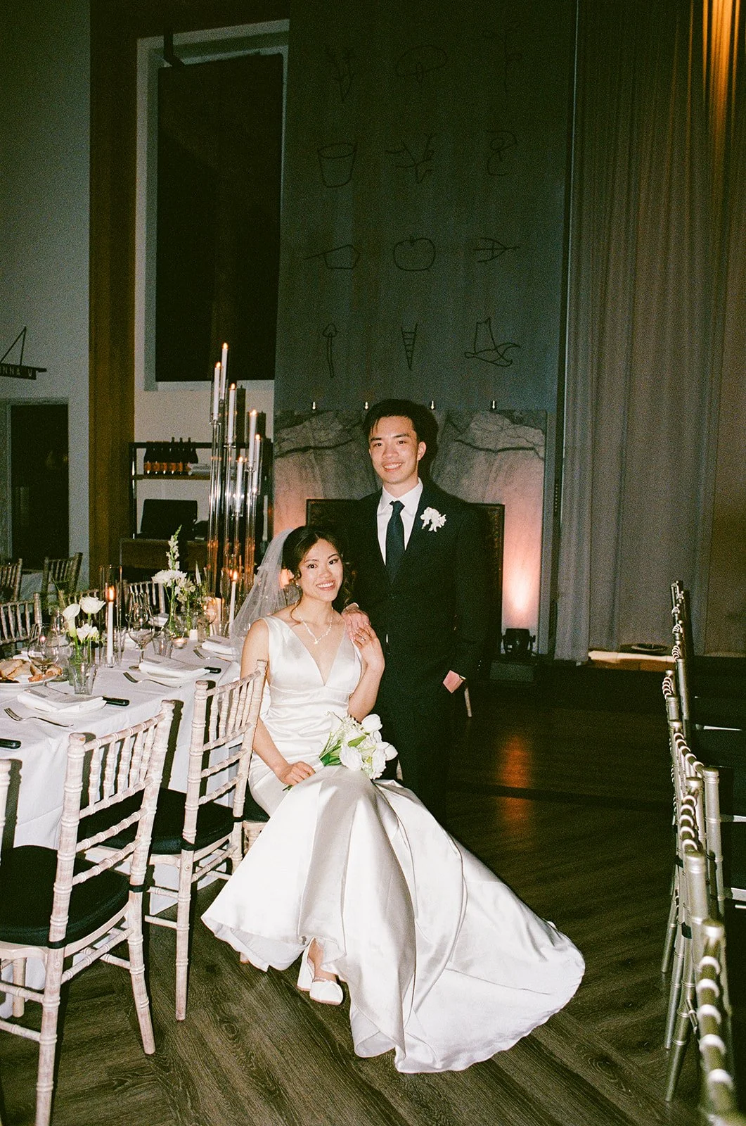 A newlywed couple at their wedding reception, with the bride seated holding a bouquet and wearing a white wedding gown, and the groom standing beside her in a black suit and tie, in a decorated banquet hall.