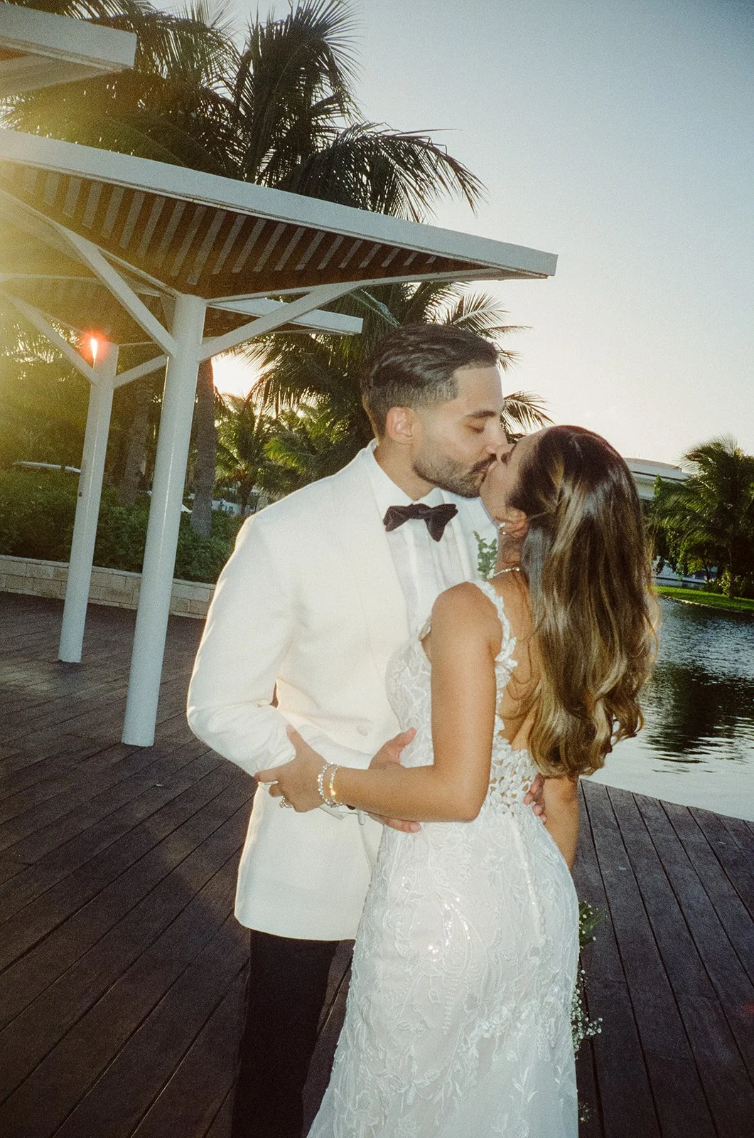 A newlywed couple sharing a kiss on a wooden dock by the water during sunset, with palm trees in the background.