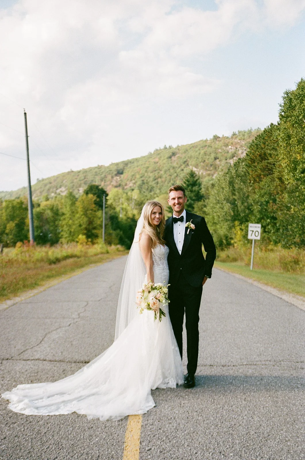 A bride and groom stand on an empty road surrounded by trees and hills, smiling. The bride wears a white wedding gown with a veil, carrying a bouquet. The groom is in a black tuxedo with a bow tie and boutonniere.