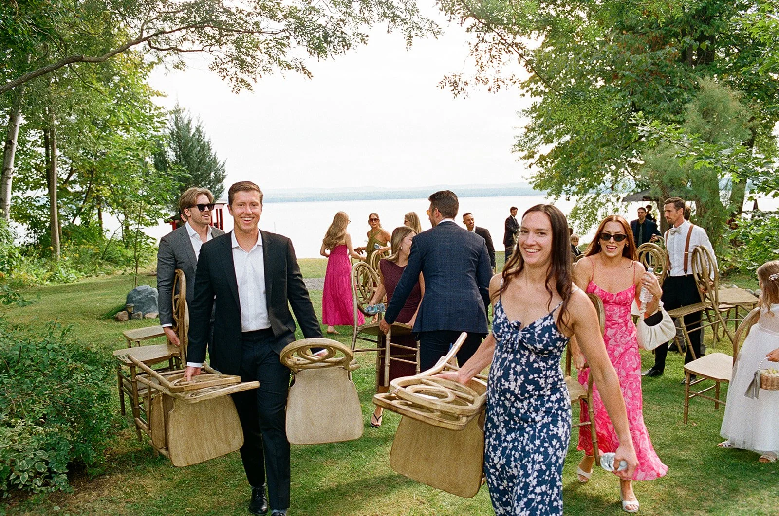People attending an outdoor event near a lake, carrying chairs and mingling on a grassy area under trees.