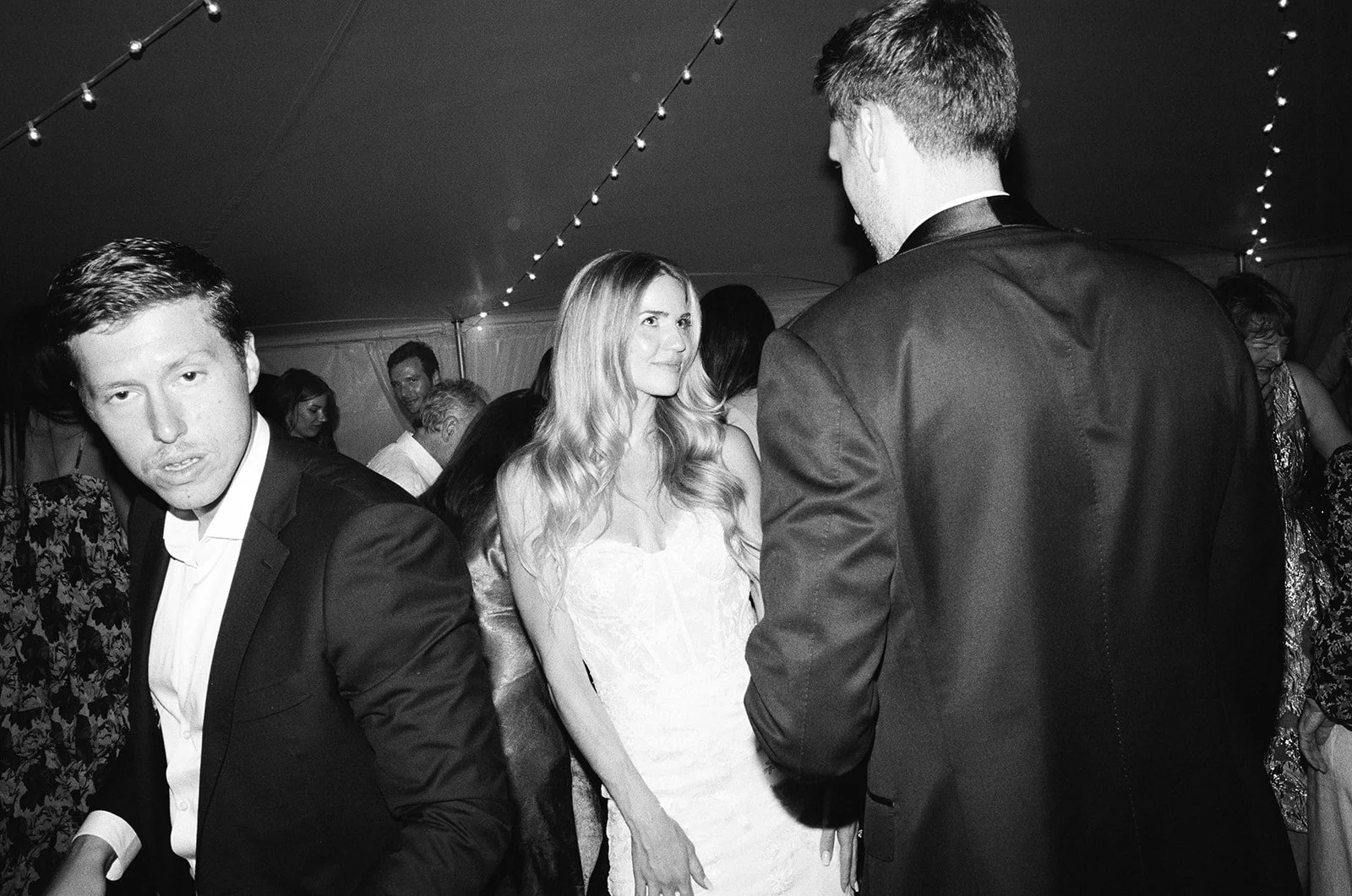 A black and white photo of a social gathering, where a woman in a white dress is engaged in conversation with a man in a tuxedo. Other guests are visible in the background under string lights.