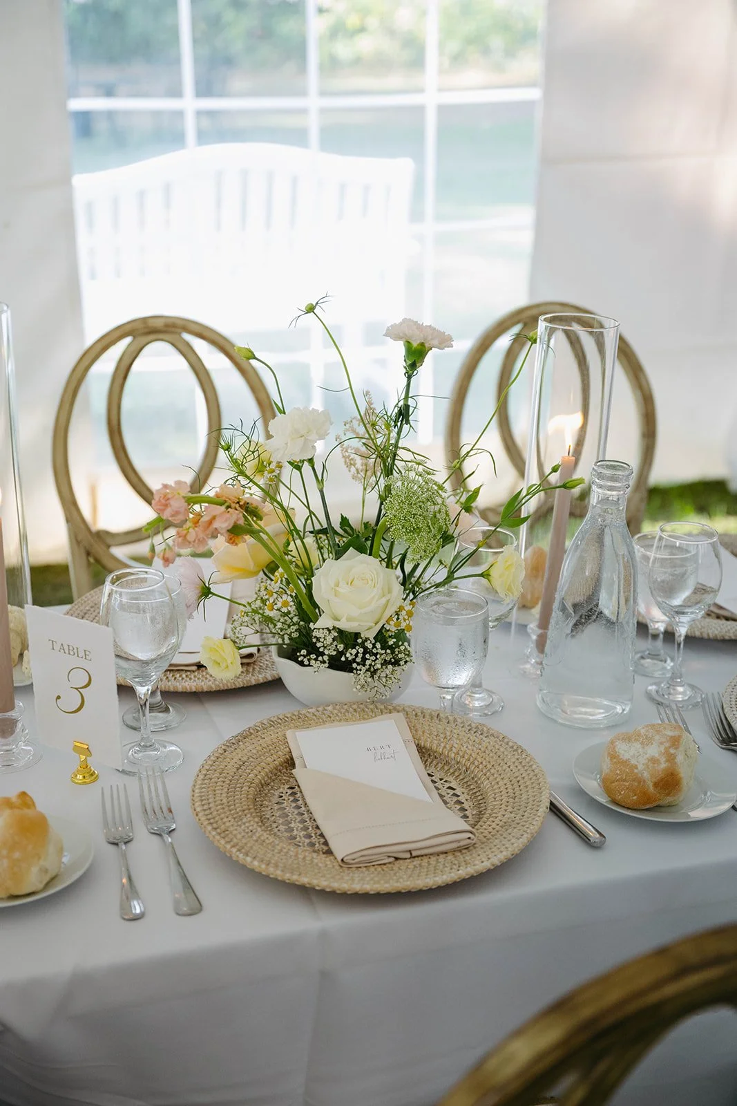 A decorated table with a floral centerpiece of white roses and greenery, set with glassware, napkins, and plates, for a formal event or wedding reception.