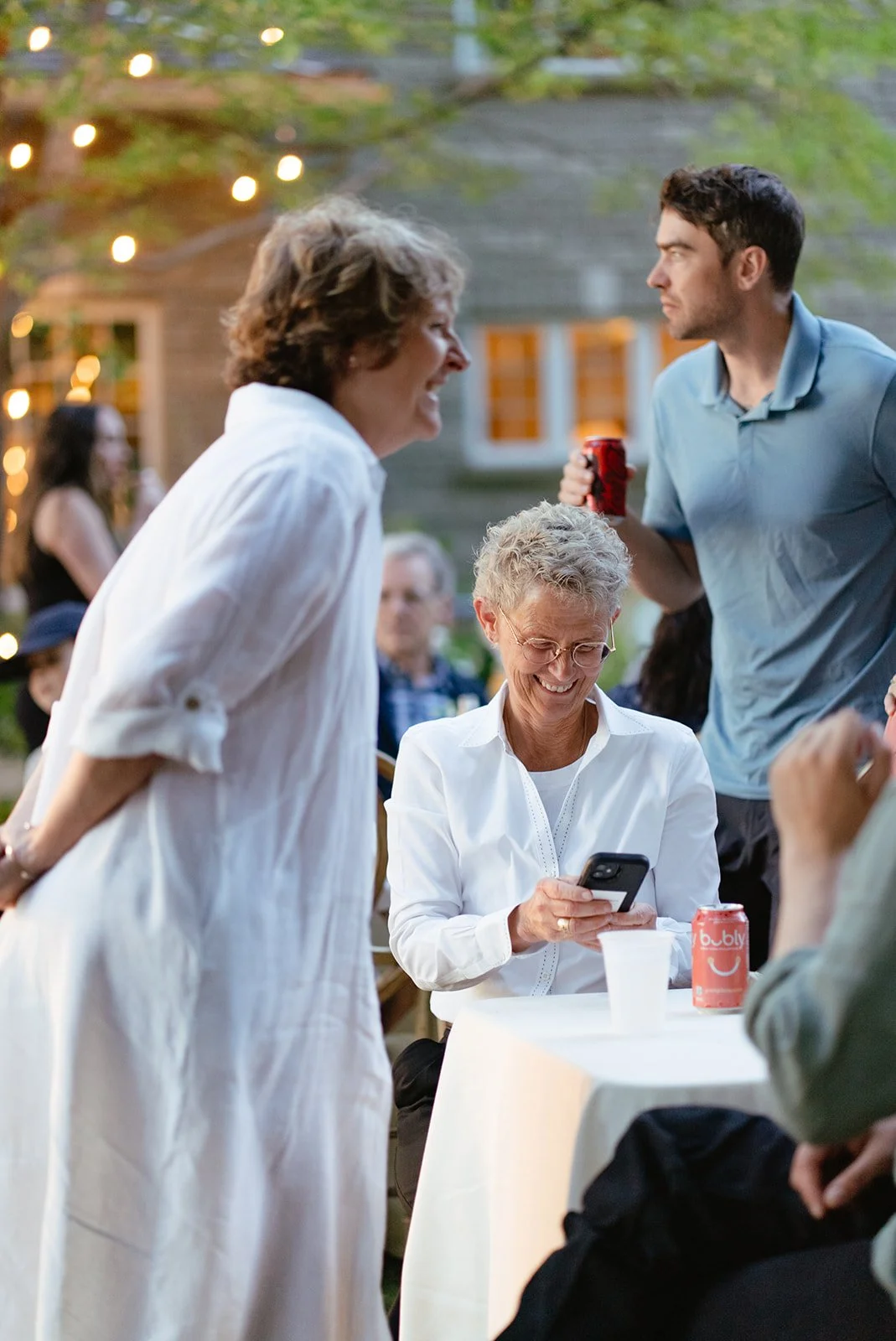 Group of people at outdoor gathering, smiling and talking, with string lights visible in the background.