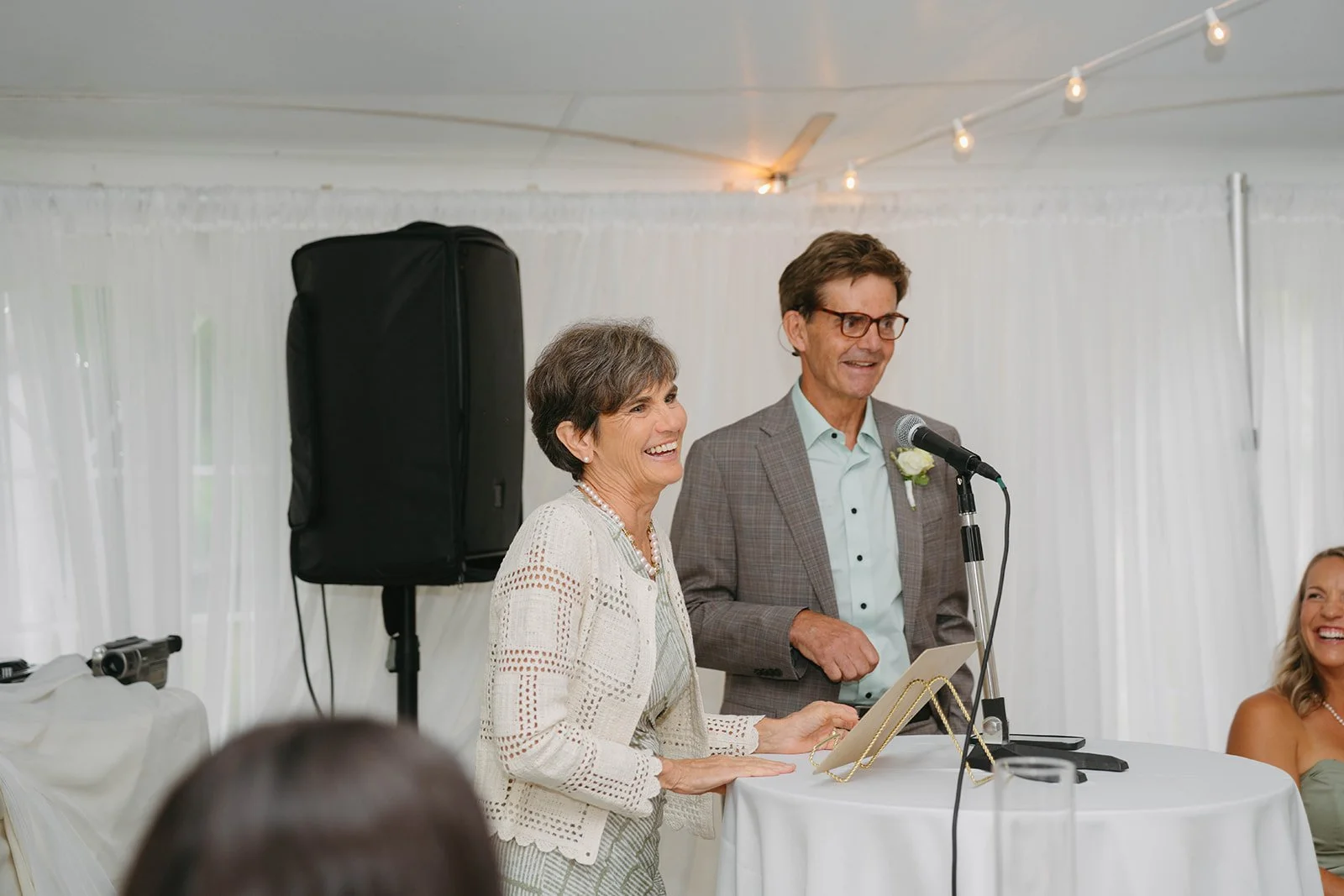 An elderly woman giving a speech at a wedding reception, smiling, standing next to a man in a gray plaid suit with a boutonnière, speaking into a microphone, in a white tent decorated with string lights.