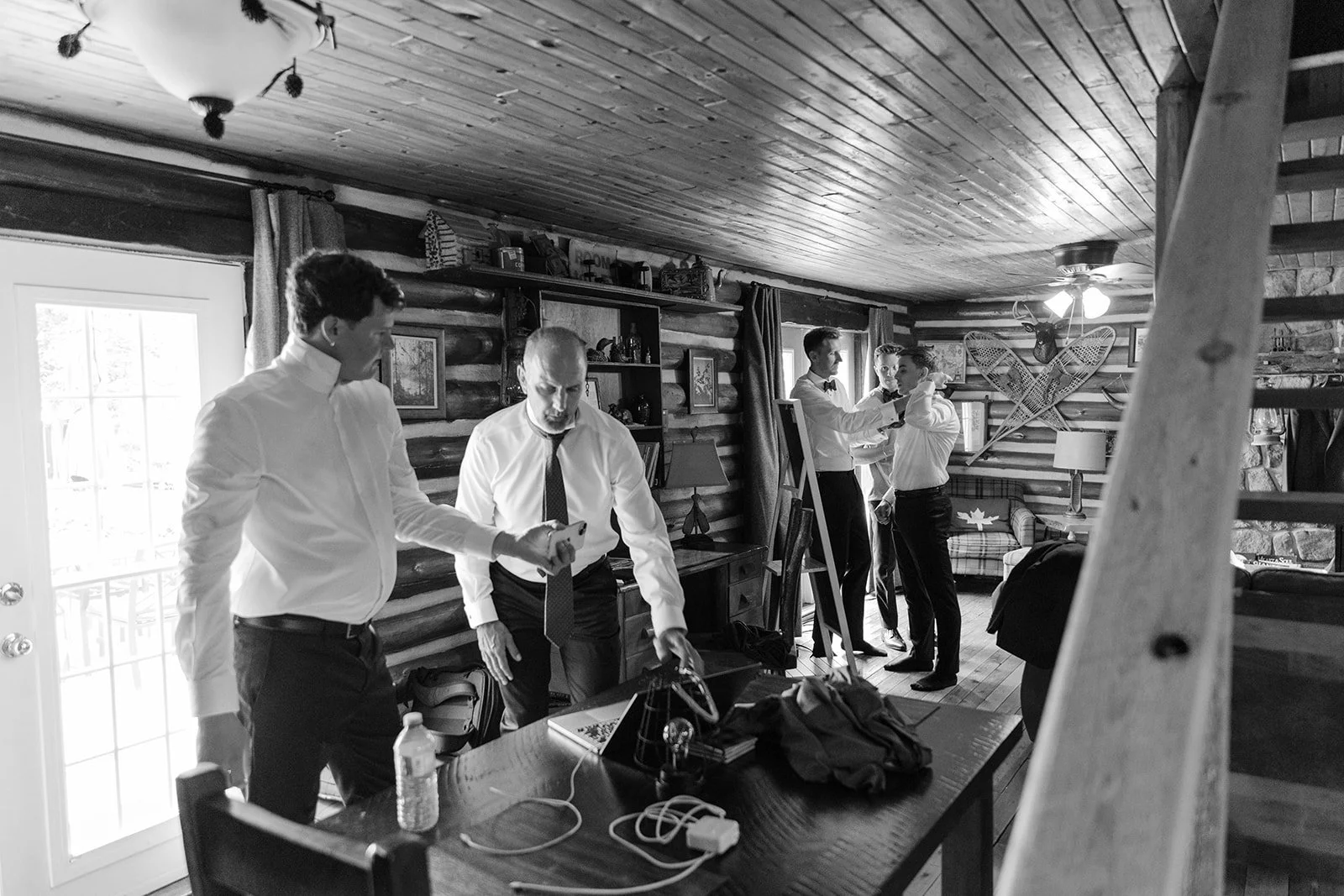 Black and white photo of five men in dress shirts and ties in a rustic log cabin with wooden walls, some of them adjusting a bowtie, standing near a bookshelf and framed pictures. A large light fixture hangs from the ceiling, and snowshoes are mounte