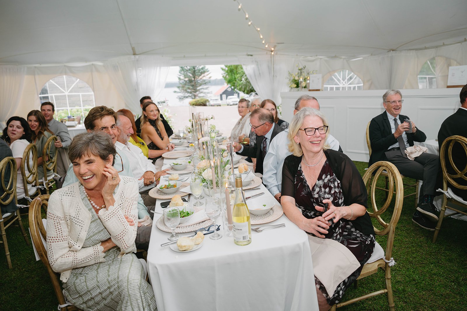 Guests seated at a long dinner table inside a white event tent, smiling and laughing, with a view of the outdoors in the background.