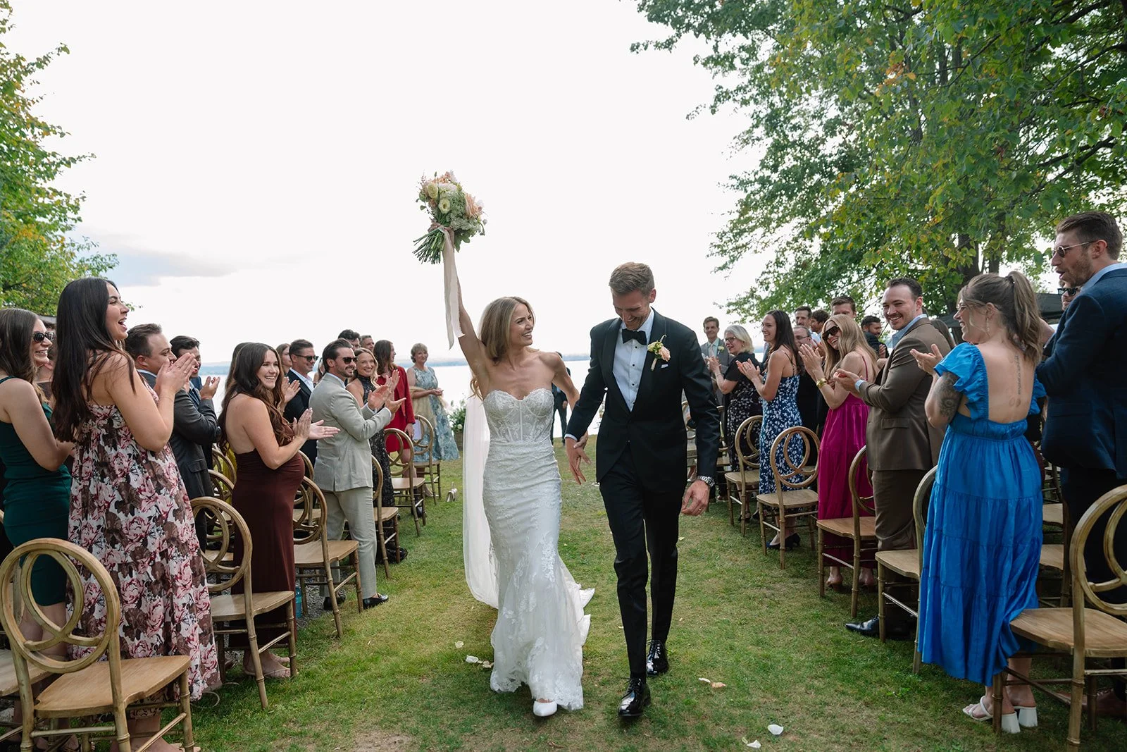 Bride and groom walking down the aisle at an outdoor wedding ceremony, surrounded by guests clapping and smiling, with a lake and trees in the background.