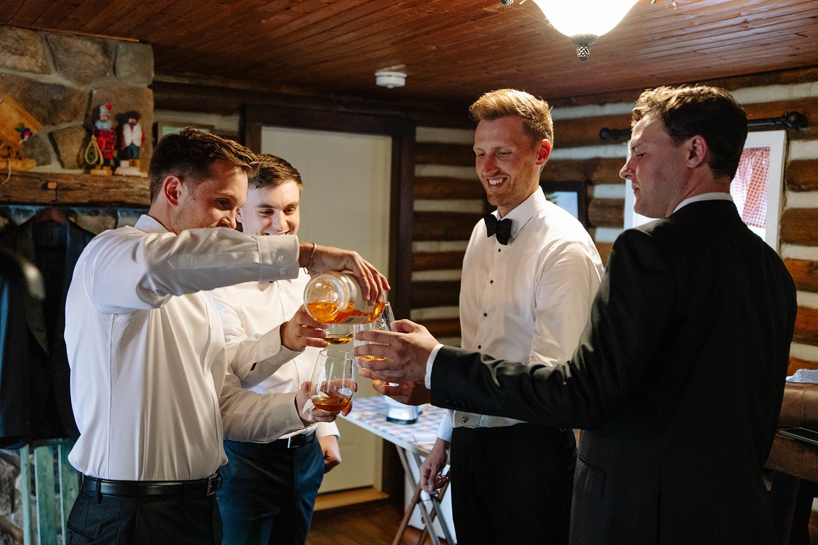 A group of five men in tuxedos and dress shirts celebrating, with one pouring a drink into another's glass, inside a log cabin style room.