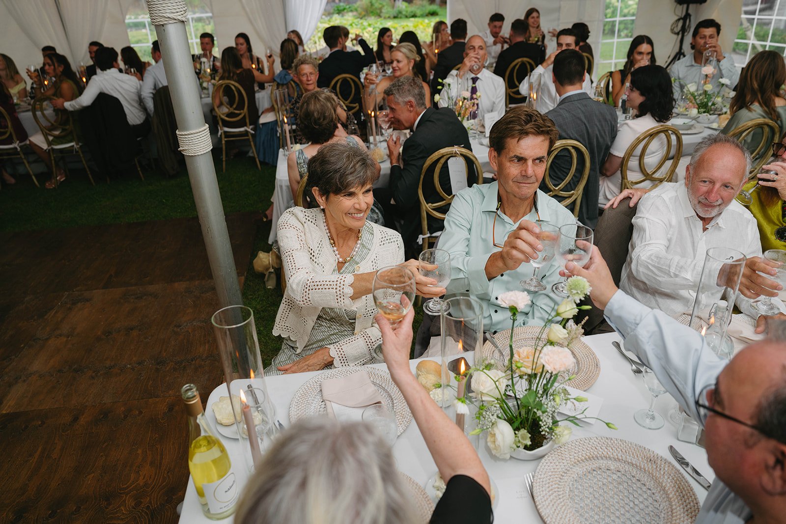A group of elderly people at a wedding reception toast their drinks at a decorated table with flowers and candles, inside a tent with other guests seated in the background.