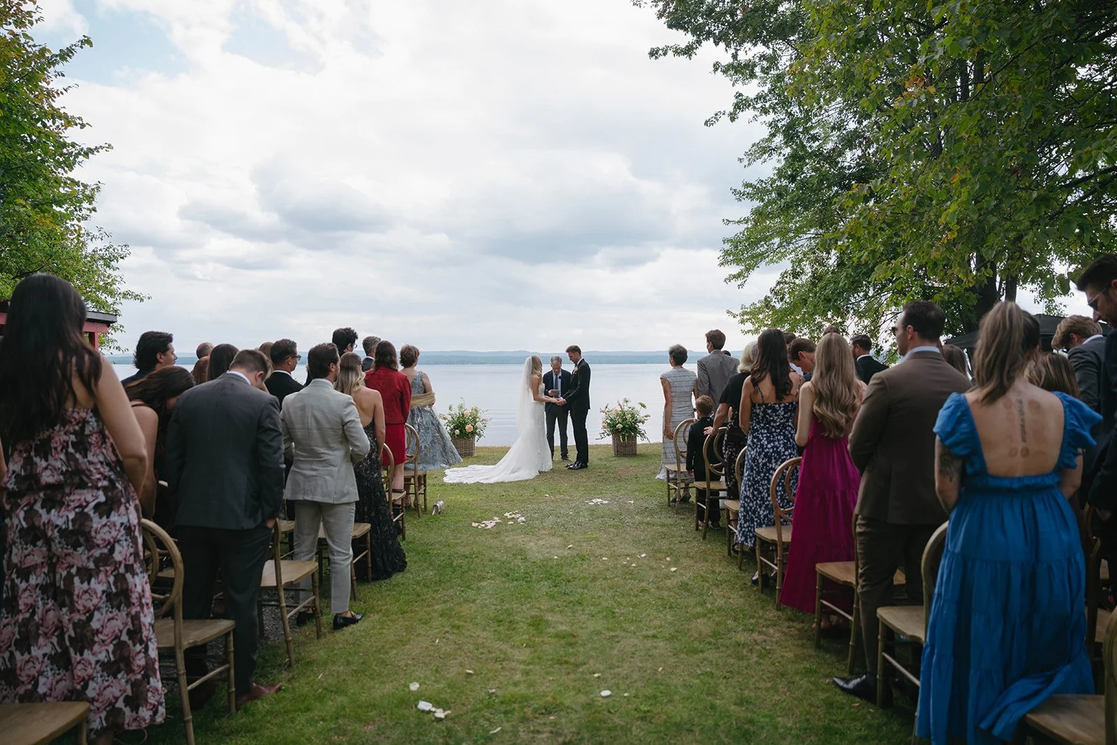A wedding ceremony taking place outdoors by a lake, with the bride and groom holding hands and an officiant in front of them. Guests are standing and seated on sides, watching the ceremony under a cloudy sky, with trees and floral arrangements nearby