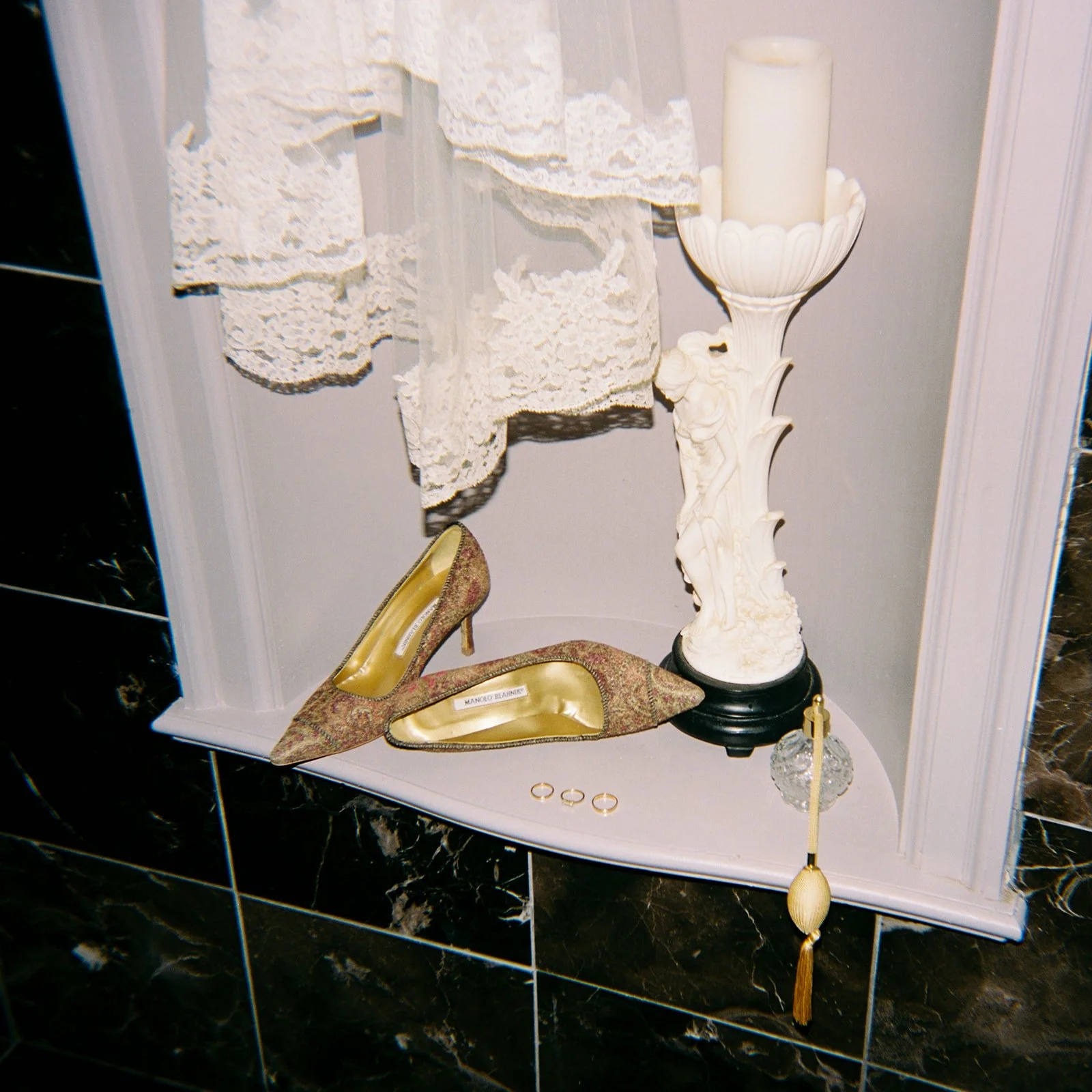 Velvet high-heeled shoes, vintage lace fabric, ornate white candle holder with candle, small glass decorative item, gold and pearl tassel, placed on a shelf with black marble wall behind.