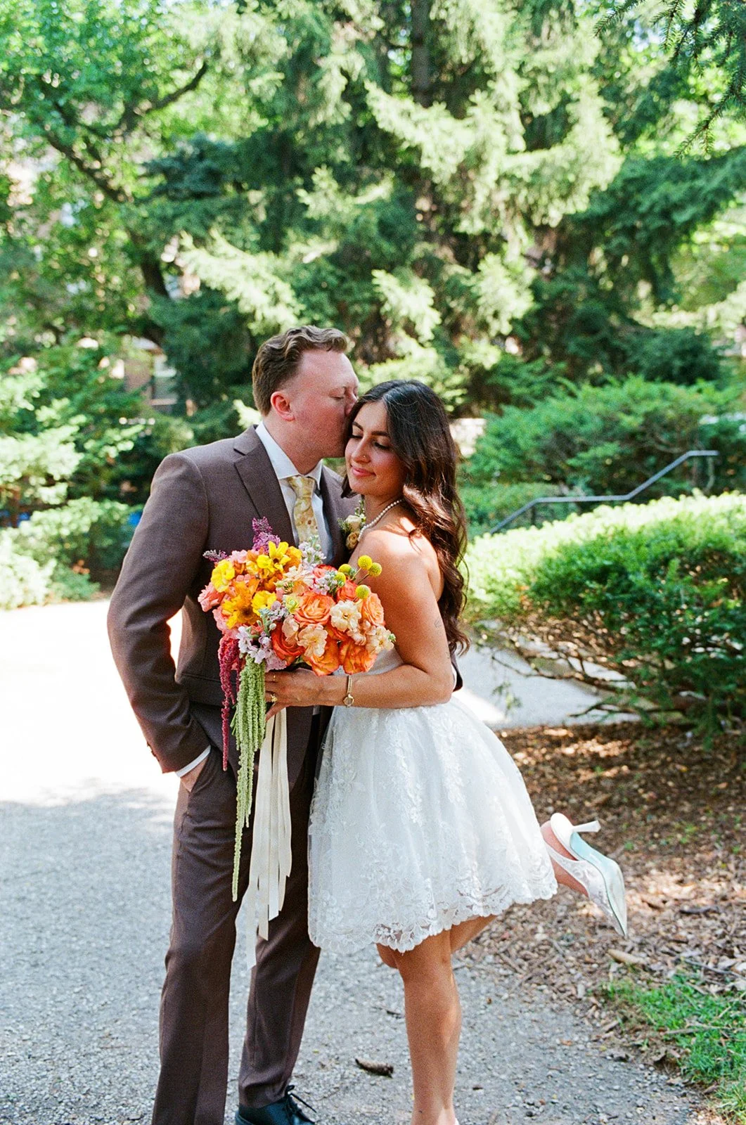 A newlywed couple stands outdoors surrounded by greenery. The groom is kissing the bride on the forehead, who is holding a colorful bouquet of flowers. The bride is wearing a white lace dress and white high heels, with her right leg lifted slightly b