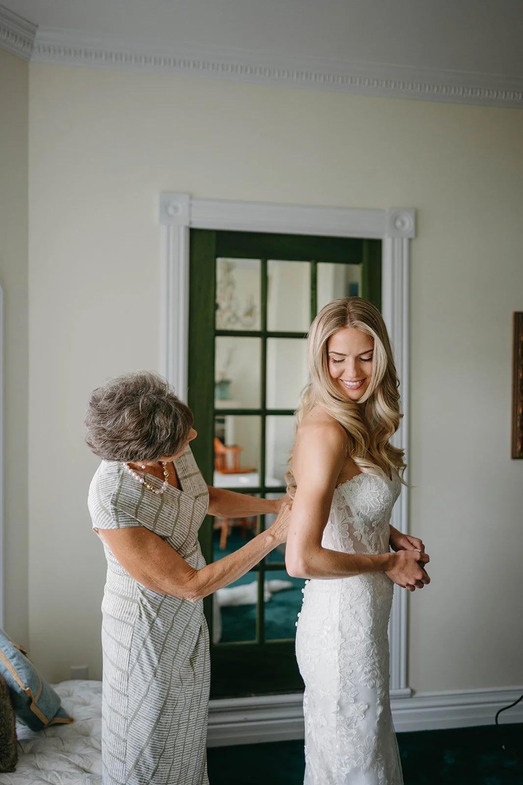 A bride in a lace wedding dress smiling as an older woman fastens the back of her dress in a room with green door and white walls.