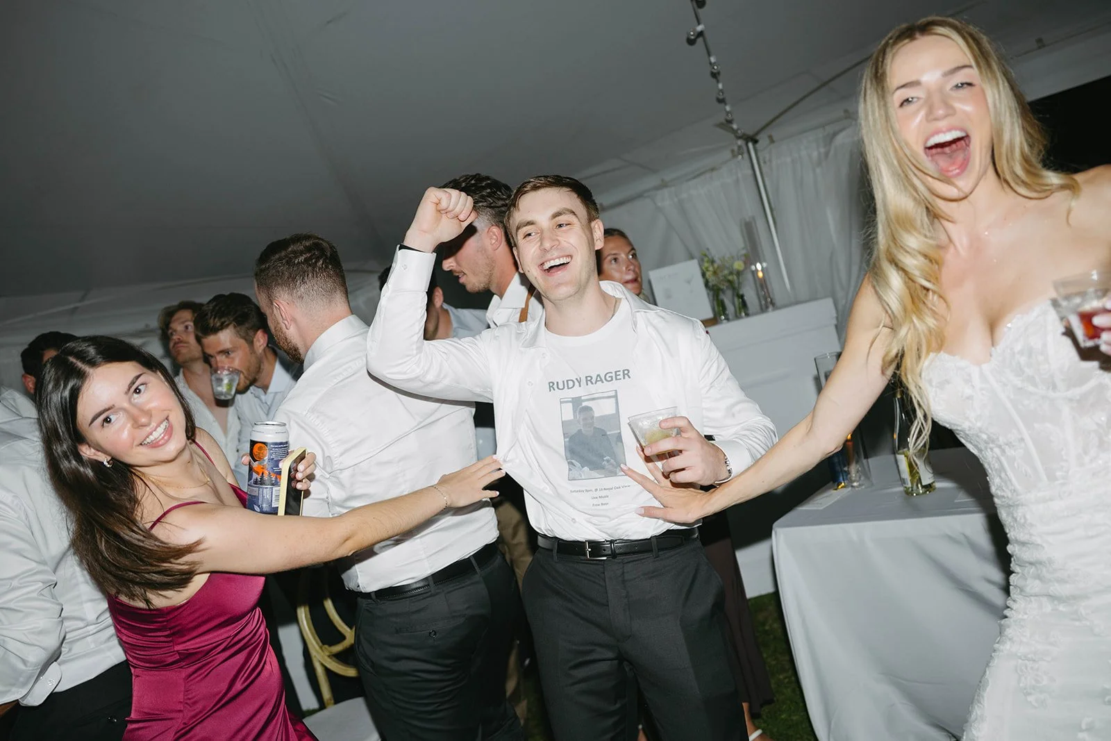 Group of people dancing and celebrating at a party, smiling and holding drinks, with a woman in a pink dress and a woman in a white dress visible.