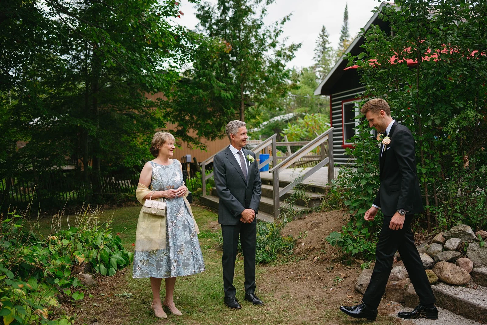 A wedding scene with three people outdoors: a woman in a floral dress and beige jacket, a man in a suit, and a groom in a black tuxedo. They are standing on a grassy area near a garden and a wooden staircase, surrounded by trees and a black house wit
