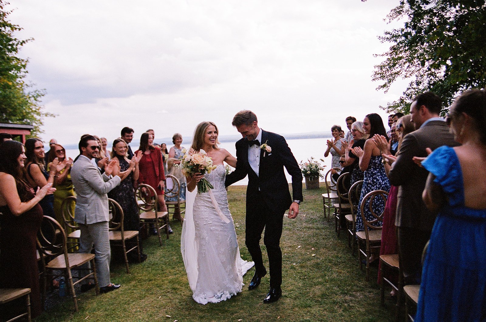 Bride and groom walking down the aisle at their outdoor wedding reception by a lake, surrounded by clapping guests.