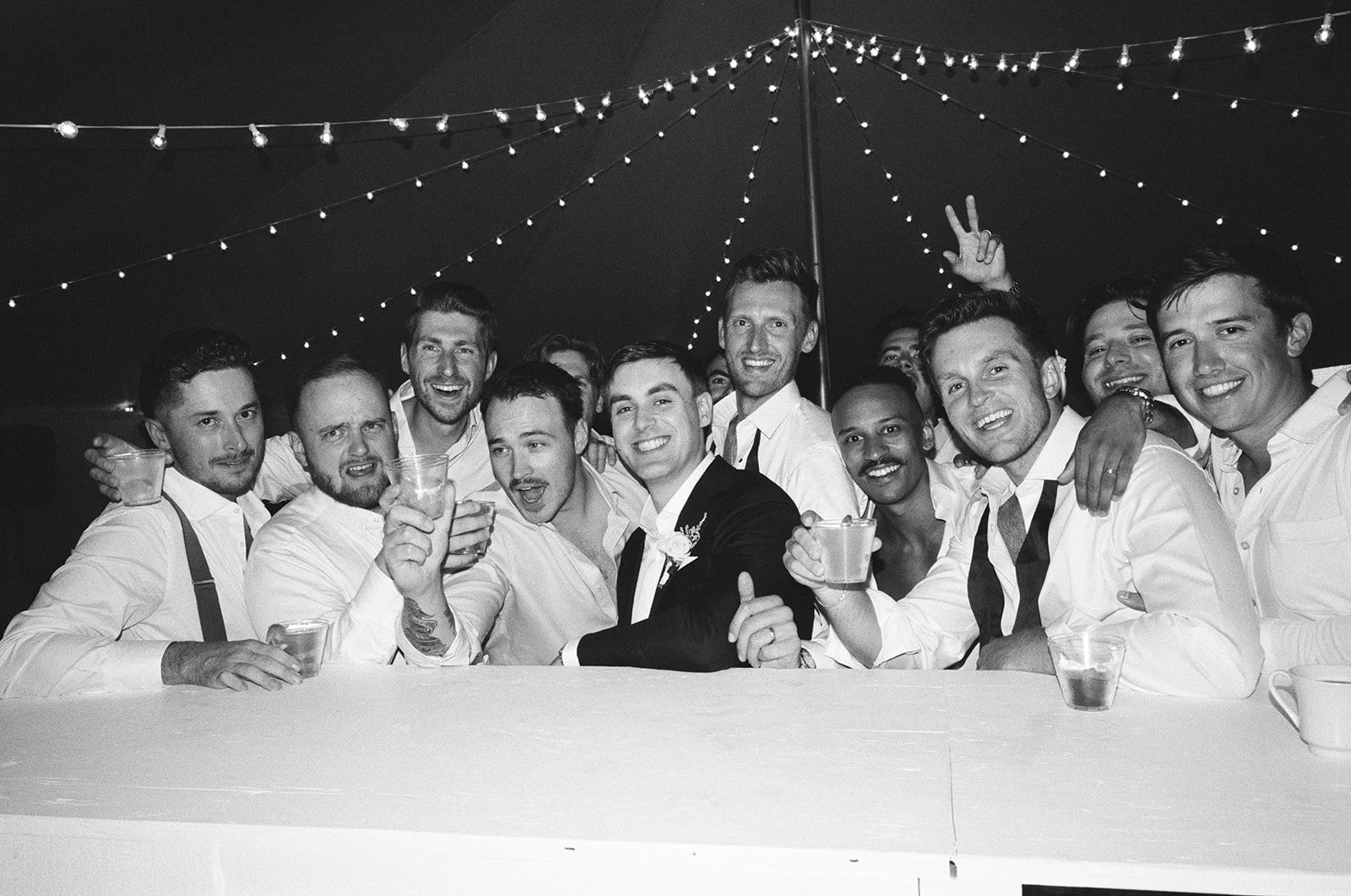 Black and white photo of a group of happy men at a celebration, some holding drinks and making peace signs, under string lights.