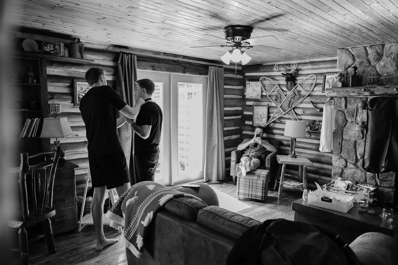Three young men in a rustic cabin living room; two are standing near a window, one is sitting on a plaid armchair taking a photo, and the third is lying on a sofa. The room has wooden walls, a stone fireplace, and vintage decor including cross-countr
