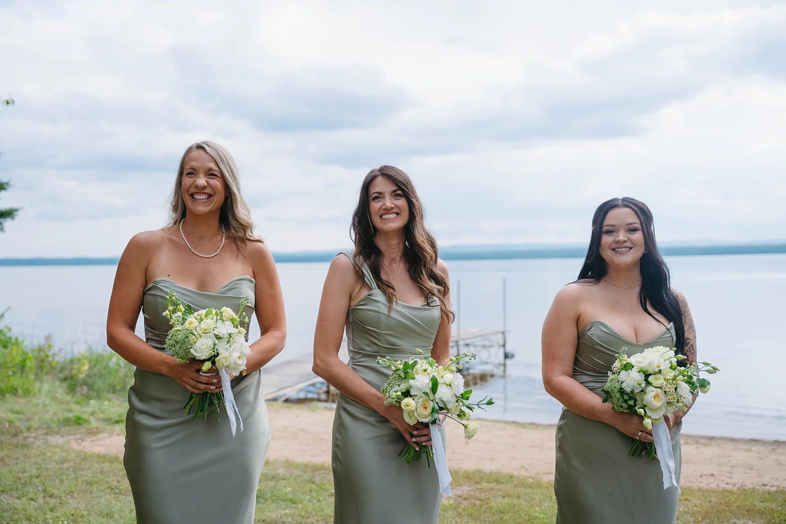 Three women in matching grey bridesmaid dresses holding bouquets, standing outdoors near a lake with a cloudy sky.