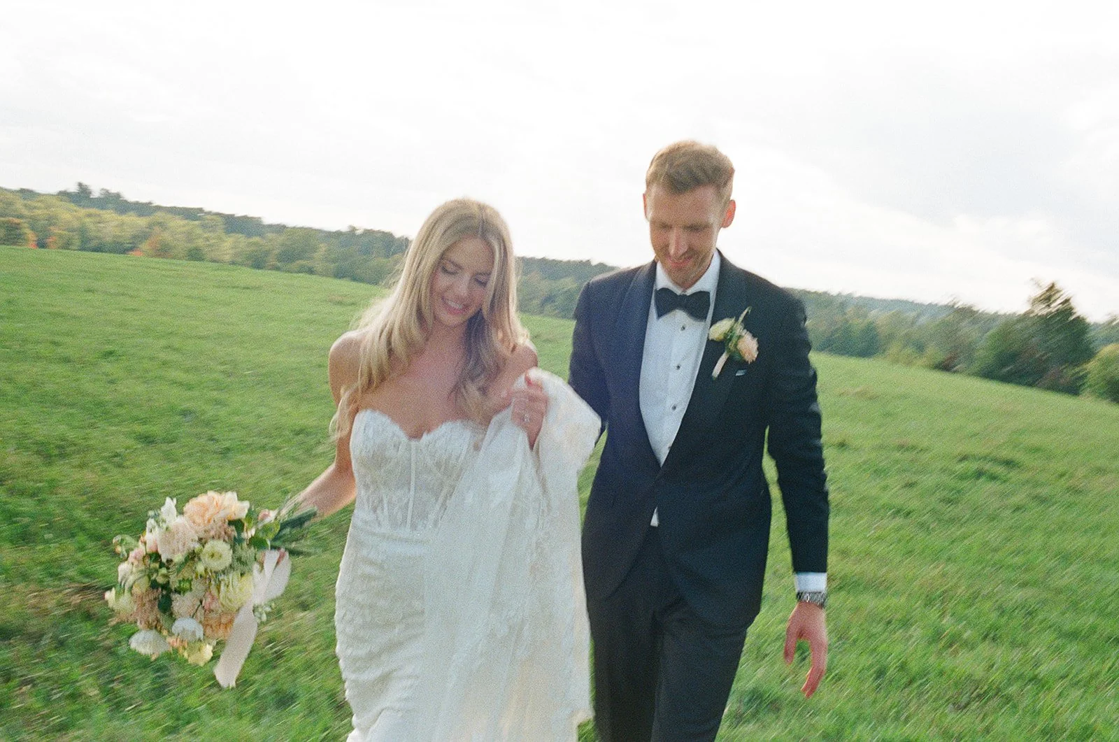 A bride and groom walking through a green field on their wedding day, smiling, with the bride holding a bouquet of flowers.