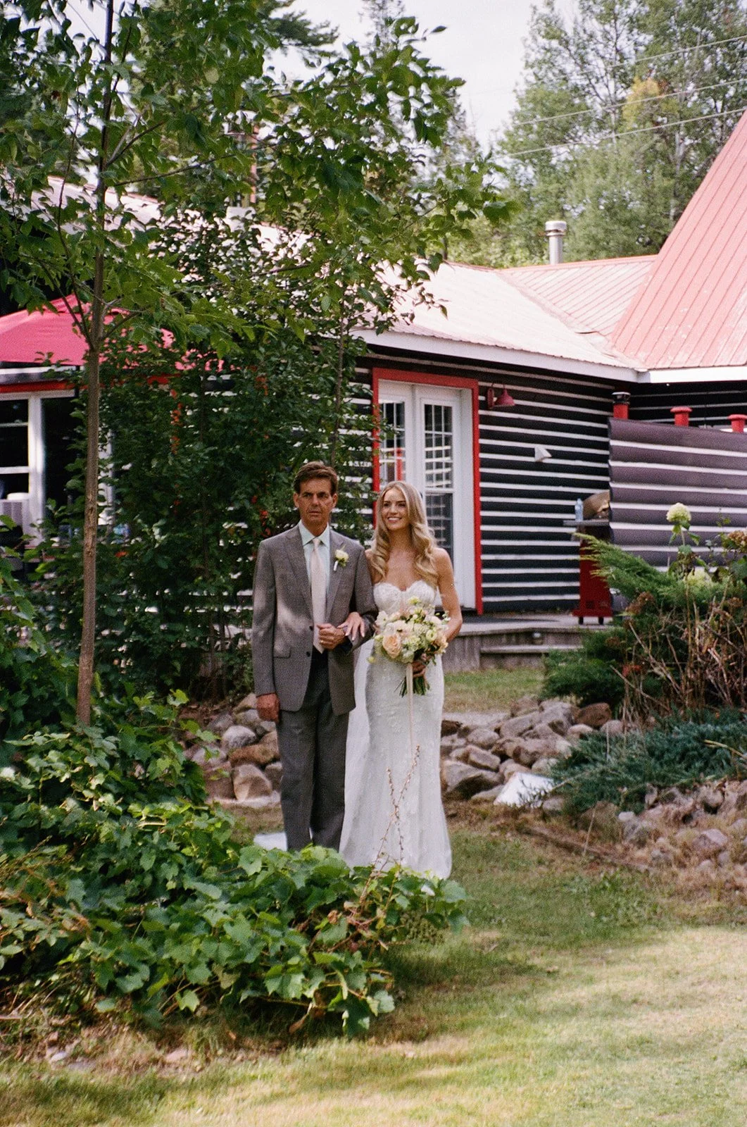 A bride in a white wedding dress holding a bouquet of flowers, walking down the aisle with a man in a suit, outdoors in front of a dark-colored house with red trim.