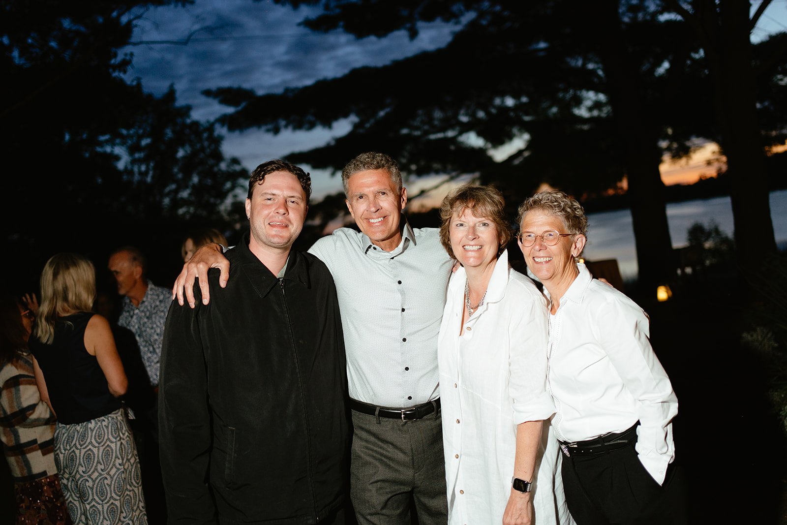 Group of five adults smiling and posing outdoors at sunset, with trees and a river in the background.