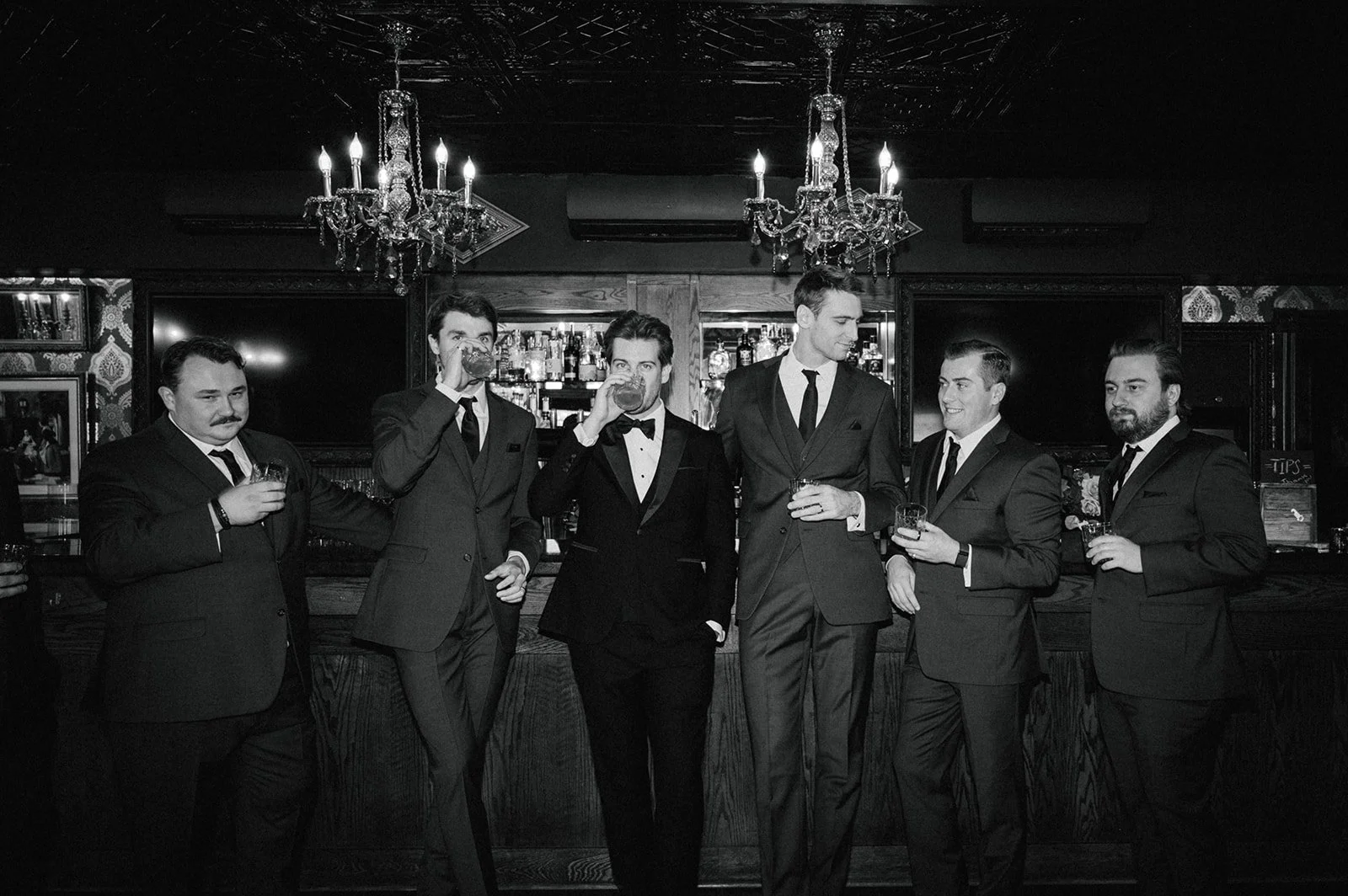 Seven men in formal suits and ties standing at a bar, drinking whiskey, in a dimly lit bar with chandeliers and artwork on the walls, black and white photograph.